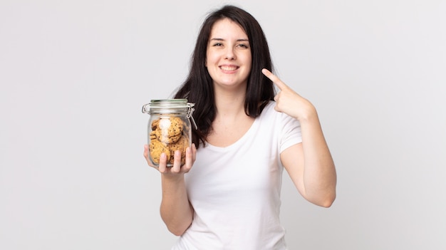 Mulher bonita sorrindo com confiança apontando para o próprio sorriso largo e segurando uma garrafa de vidro de biscoitos