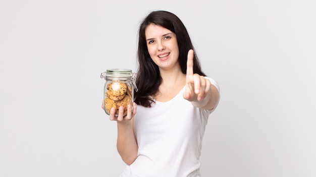 Mulher bonita sorrindo com orgulho e confiança fazendo o número um e segurando uma garrafa de vidro de biscoitos