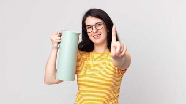 Mulher bonita sorrindo com orgulho e confiança fazendo o número um e segurando uma garrafa térmica de café