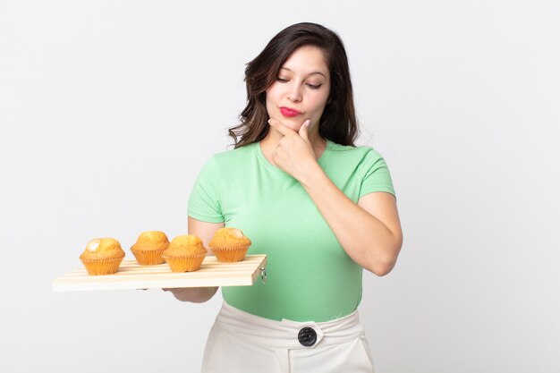 Mulher bonita sorrindo com uma expressão feliz e confiante, com a mão no queixo e segurando uma bandeja de muffins