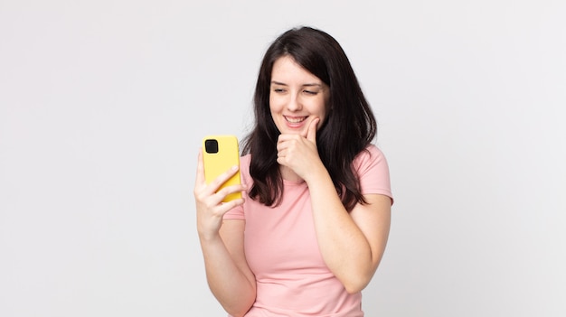 Mulher bonita sorrindo com uma expressão feliz e confiante com a mão no queixo usando um telefone inteligente