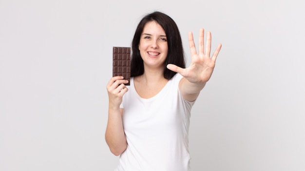 Mulher bonita sorrindo e parecendo amigável, mostrando o número cinco e segurando uma barra de chocolate