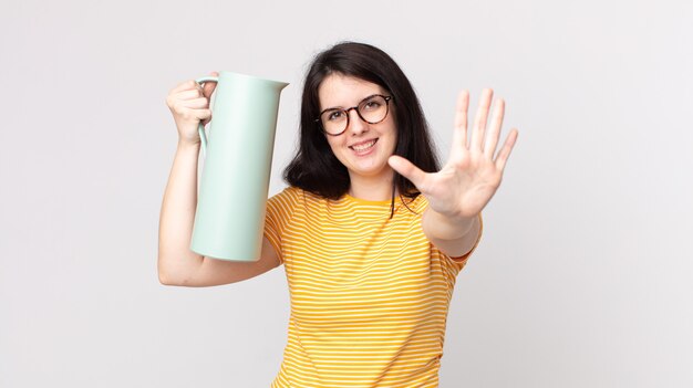 Mulher bonita sorrindo e parecendo amigável, mostrando o número cinco e segurando uma garrafa térmica de café