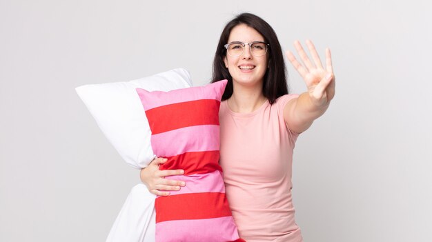 Mulher bonita sorrindo e parecendo amigável, mostrando o número quatro de pijama e segurando um travesseiro