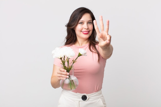 Mulher bonita sorrindo e parecendo amigável, mostrando o número três e segurando um vaso de flores decorativo