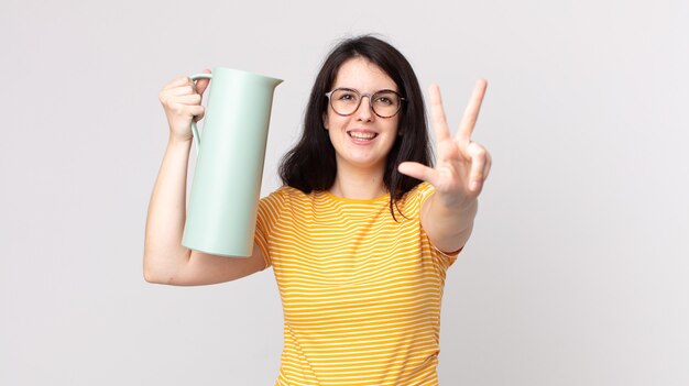 Foto mulher bonita sorrindo e parecendo amigável, mostrando o número três e segurando uma garrafa térmica de café