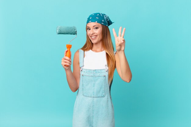 Mulher bonita sorrindo e parecendo amigável, mostrando o número três pintando uma nova parede de casa