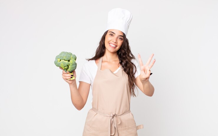 Foto mulher bonita sorrindo e parecendo amigável, mostrando o número três usando um avental e segurando um brócolis