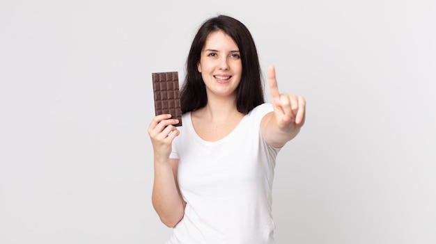 Mulher bonita sorrindo e parecendo amigável, mostrando o número um e segurando uma barra de chocolate