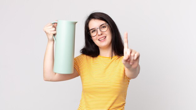 Mulher bonita sorrindo e parecendo amigável, mostrando o número um e segurando uma garrafa térmica de café