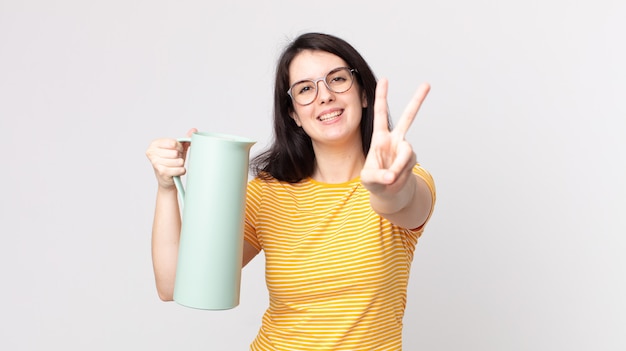 Mulher bonita sorrindo e parecendo feliz, gesticulando vitória ou paz e segurando uma garrafa térmica de café