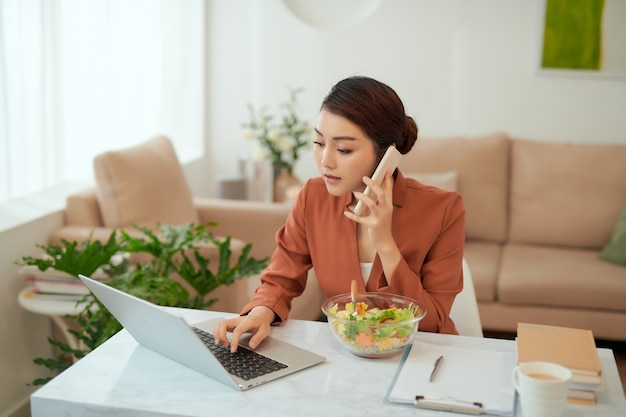 Mulher bonita sorrindo enquanto trabalhava em casa, ao telefone com uma salada