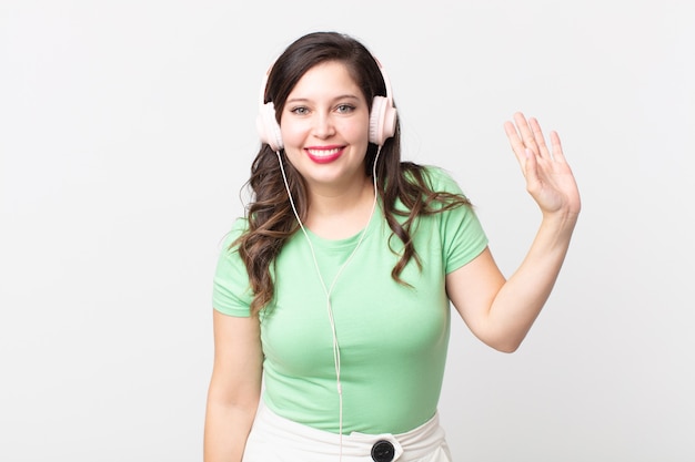 Mulher bonita sorrindo feliz, acenando com a mão, dando as boas-vindas e cumprimentando você ouvindo música com fones de ouvido