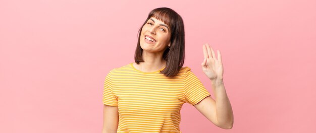 Mulher bonita sorrindo feliz e alegre, acenando com a mão, dando as boas-vindas e cumprimentando você ou dizendo adeus