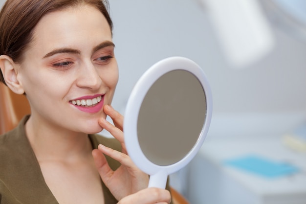 Mulher bonita sorrindo, olhando no espelho na clínica odontológica