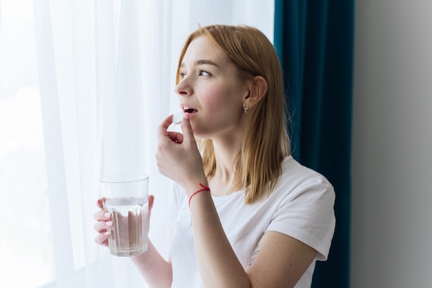 Mulher bonita toma um comprimido perto da janela com um copo d'água. bela mulher sorridente tomando comprimido de vitamina. suplemento alimentar.