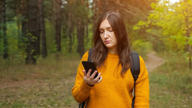 Mulher bonita turista com mochila em um elegante suéter laranja olha para a tela do novo celular caminhando pela estrada na floresta verde, luz do sol