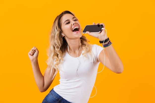 Mulher bonita usando fones de ouvido cantando enquanto segura o celular como microfone, isolado na parede amarela