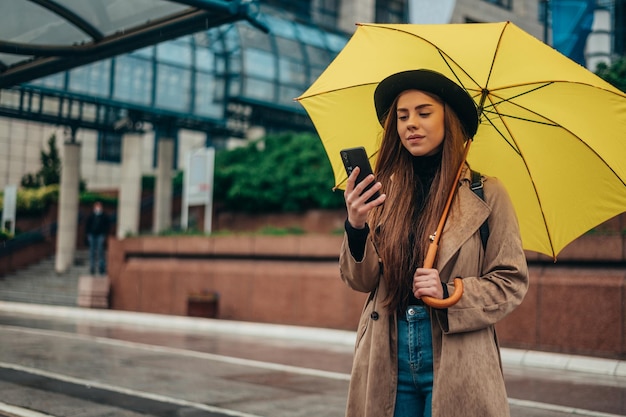 Mulher bonita usando um smartphone e segurando um guarda-chuva amarelo enquanto caminhava na cidade em um dia chuvoso