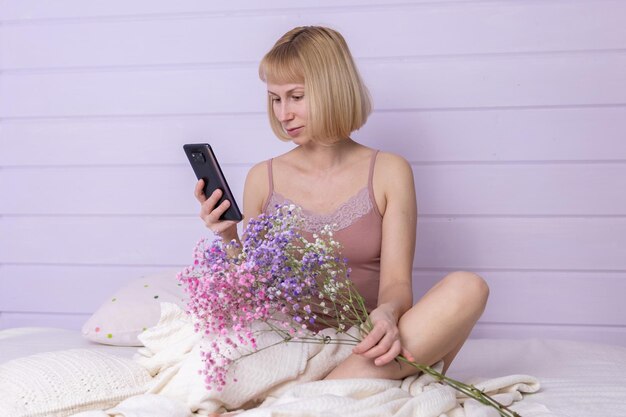 Mulher bonita vestindo lingerie de luxo, sentado na cama em linho branco em apartamento aconchegante e usando smartphone Mulher sexy está sentada com o telefone na cama no foco suave do quarto