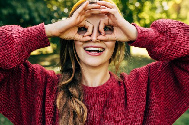 Mulher bonita vestindo um suéter vermelho fazendo gesto okey como binóculos sorrindo amplamente e olhando por entre os dedos muito fingir que olha através de binóculos em algo sobre o fundo da natureza