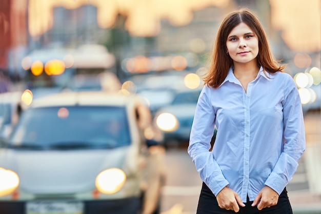 Mulher branca de 26 anos de camisa em pé perto do tráfego na rua iluminada por faróis de carros em noite de verão.