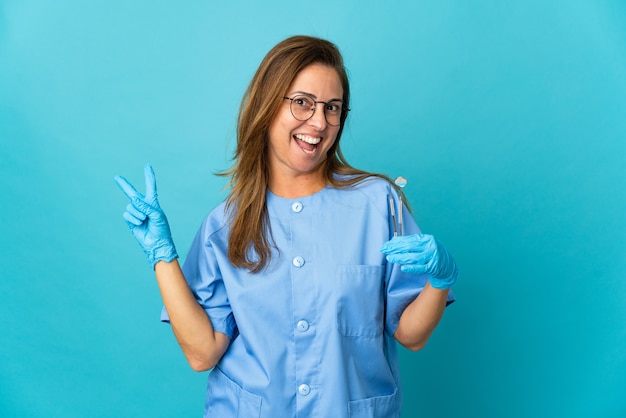 Mulher brasileira de meia-idade, dentista, segurando ferramentas sobre uma parede isolada, sorrindo e mostrando o sinal da vitória