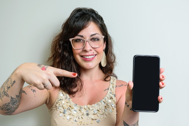 Foto mulher brasileira de óculos, tatuada, apontando o dedo e mostrando o smartphone.