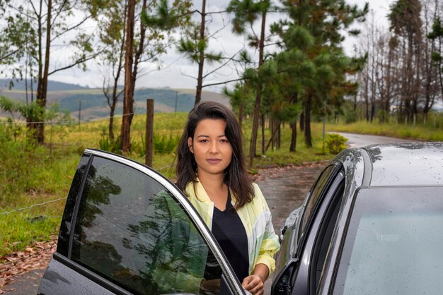 Mulher brasileira entrando no carro e árvores ao fundo em dia chuvoso