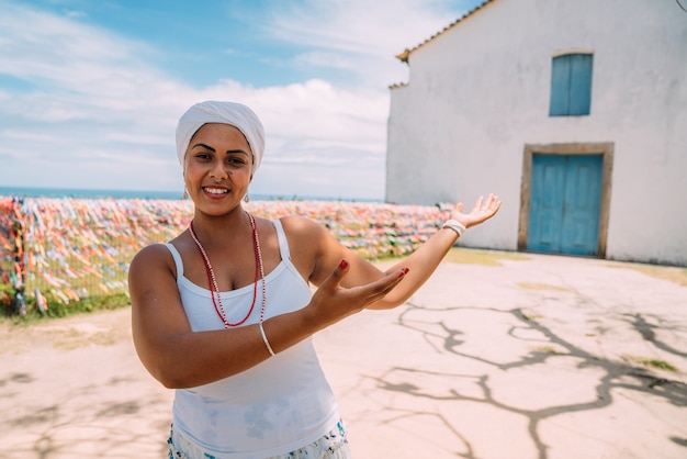 Mulher brasileira feliz vestida com o tradicional traje baiano mostrando algo na palma da mão, olha para a câmera, com o centro histórico de porto seguro ao fundo