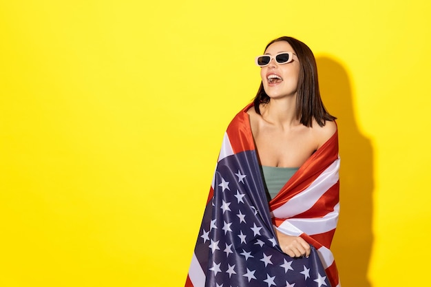 Mulher bronzeada encantadora em um maiô em um fundo amarelo Um modelo com a bandeira estrelada dos EUA e óculos de sol posa Conceito de verão