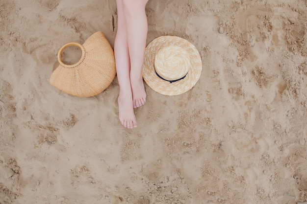 Mulher bronzeada pernas, chapéu de palha e bolsa na praia de areia. Relaxando na praia, com os pés na areia.