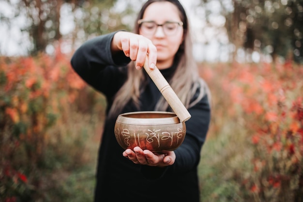 Mulher budista vestida de preto segurando e tocando uma tigela tibetana com uma vara de madeira.