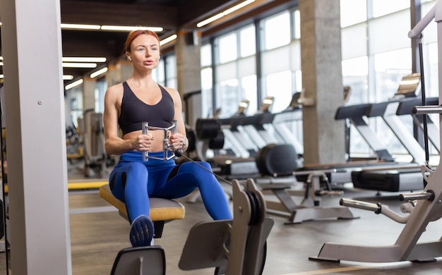 Mulher cabeluda em roupas esportivas treina os músculos das costas na máquina de exercício de cabo de remo horizontal no ginásio