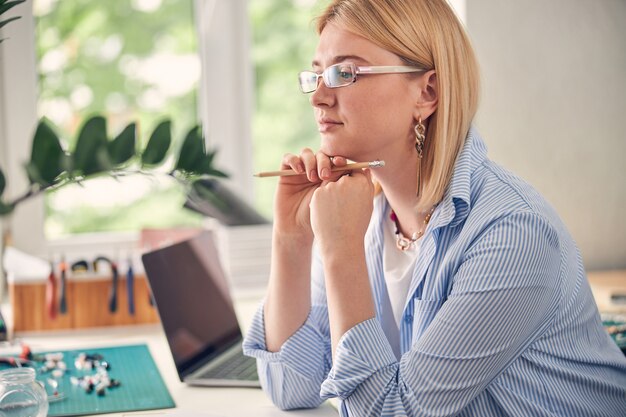 Mulher calma e inteligente mantendo as mãos embaixo do queixo enquanto descansa na mesa de trabalho