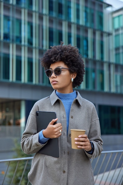 mulher caminha na rua da cidade com tablet digital e xícara de café de papel usa tecnologias modernas usa óculos de sol e jaqueta posa contra edifícios modernos