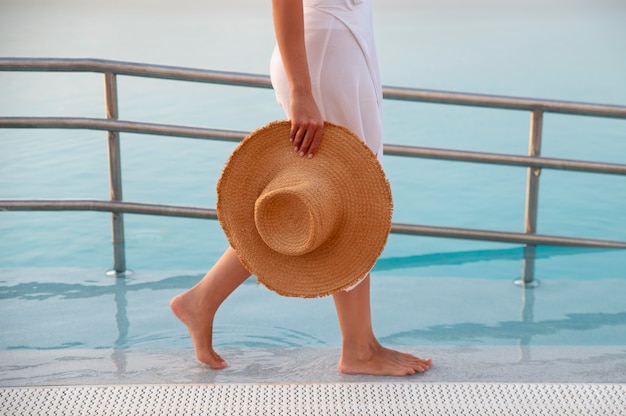 Foto mulher caminhando ao lado de uma piscina segurando seu chapéu de palha