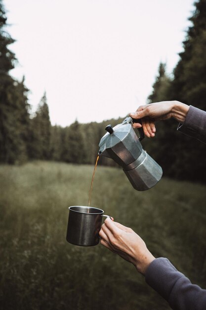 Foto mulher caminhante derramando café quente de uma chaleira de metal em uma caneca de esmalte no conceito ao ar livre da floresta