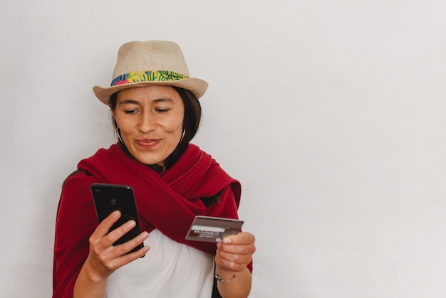 Mulher camponesa latina usando chapéu e um xale vermelho segurando um telefone celular e um cartão de crédito em suas mãos Conceito de pagamento eletrônico