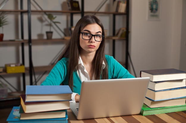 Mulher cansada de óculos com muitos livros e laptop está sentada à mesa de madeira