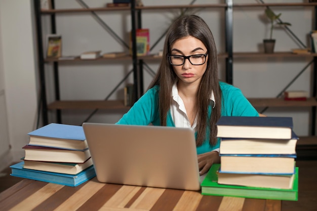 Mulher cansada de óculos com muitos livros e laptop está trabalhando na mesa