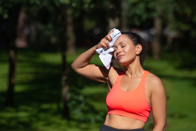 Mulher cansada e desportiva, limpe o rosto com uma toalha após o exercício produtivo ao ar livre.