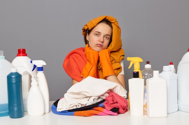 Mulher cansada entediada posando no local de trabalho com detergentes de limpeza isolados sobre fundo cinza olhando para a câmera com rosto infeliz sentindo fadiga