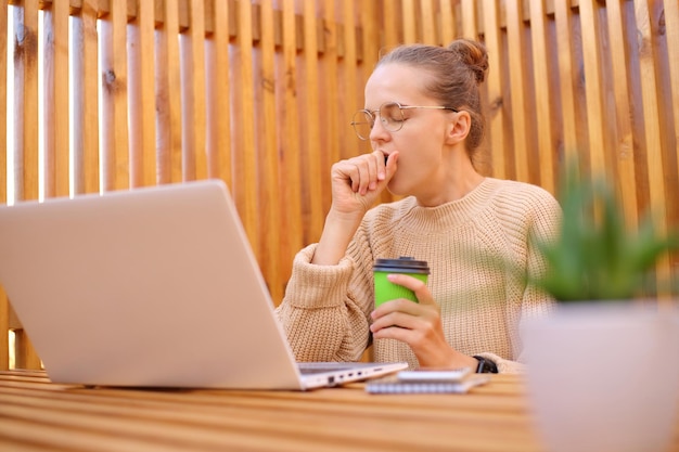 Mulher cansada freelance vestindo suéter bege sentado em um café ao ar livre trabalhando em um trabalho remoto de laptop sentindo fadiga bocejando parece sonolento