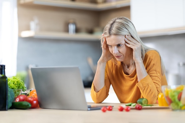 Mulher cansada olhando para a tela do laptop enquanto cozinha em casa