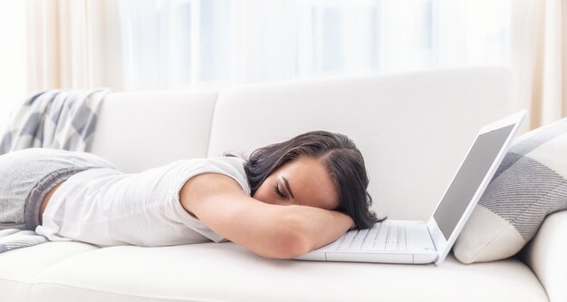 Mulher cansada que adormeceu em seu laptop deitada em um sofá em um ambiente branco de uma sala de estar.
