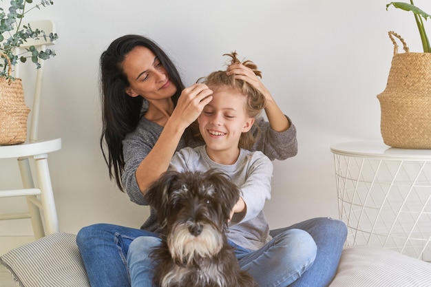 Mulher carinhosa fazendo penteado para menino otimista enquanto está sentada no chão com um lindo cachorro Schnauzer Miniatura na sala de estar iluminada