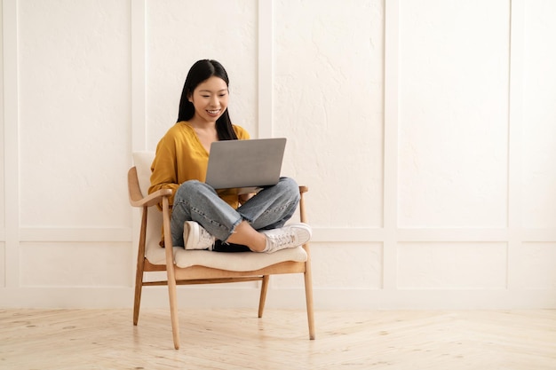 Mulher casual asiática jovem positiva sentada na poltrona usando laptop