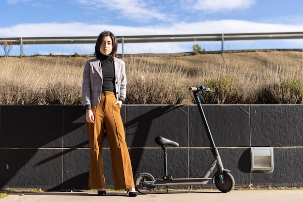 Mulher casual inteligente ao lado de uma scooter elétrica na calçada Jovem perto de uma scooter elétrica olhando para a câmera