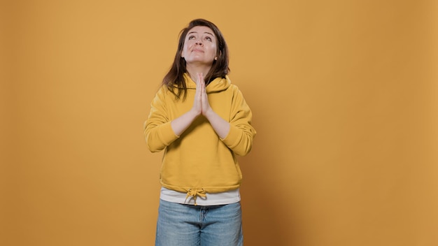 Mulher casual se sentindo preocupada, olhando para cima orando e pedindo perdão com as palmas das mãos juntas no estúdio. Jovem humilde vestindo capuz sendo religioso tendo esperança e fé.